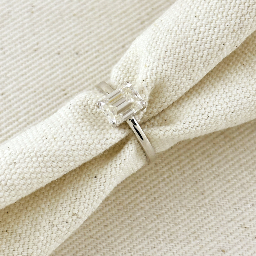 A silver ring with a clear baguette-cut gemstone, shown resting on a textured cream-colored woven fabric.