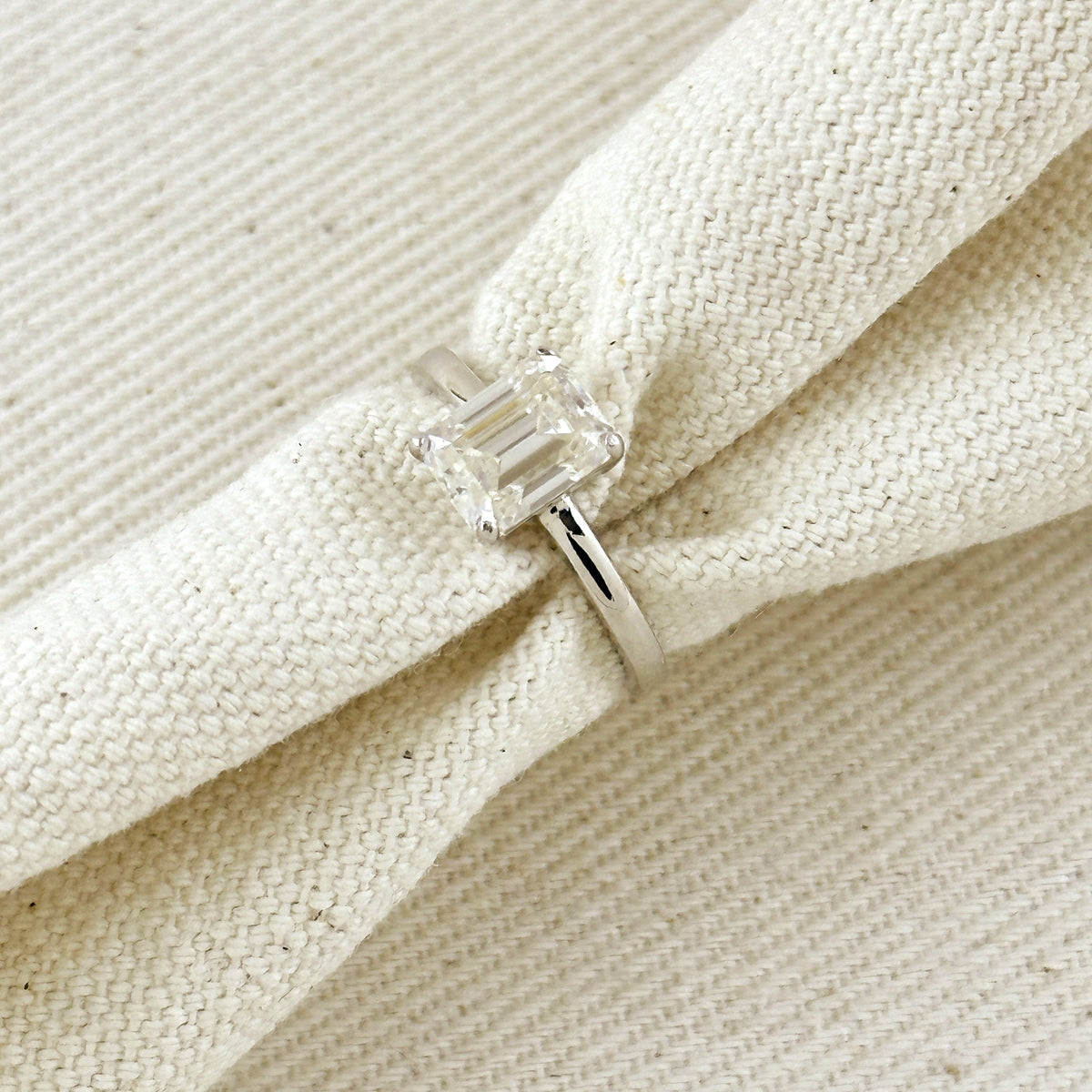 A silver ring with a clear baguette-cut gemstone, shown resting on a textured cream-colored woven fabric.