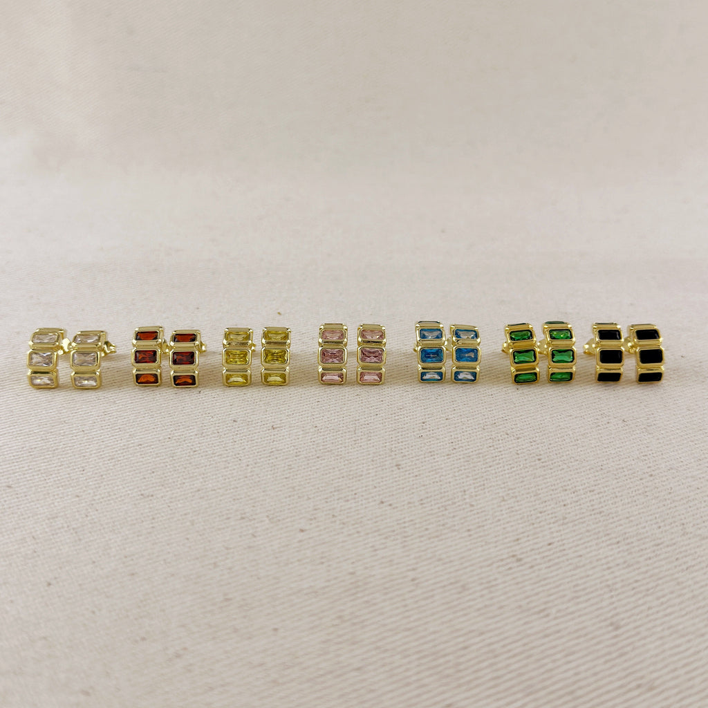 A row of nine gold-plated rectangular stud earrings with various colored rectangular gemstones, displayed on a light beige textured fabric background.