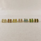 A row of nine gold-plated rectangular stud earrings with various colored rectangular gemstones, displayed on a light beige textured fabric background.