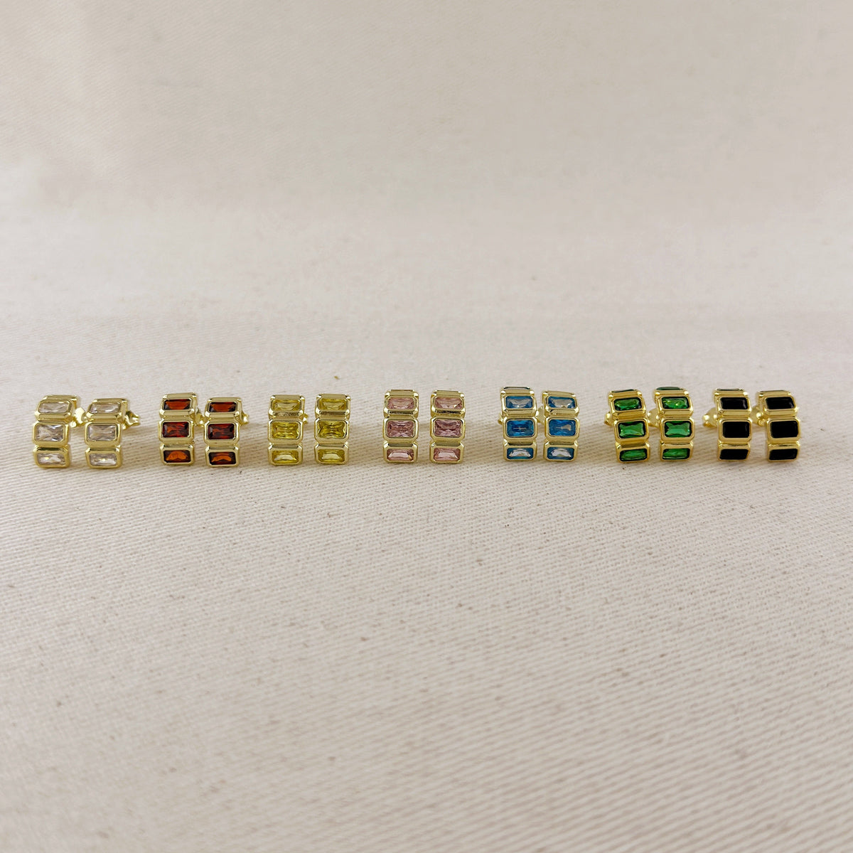 A row of nine gold-plated rectangular stud earrings with various colored rectangular gemstones, displayed on a light beige textured fabric background.